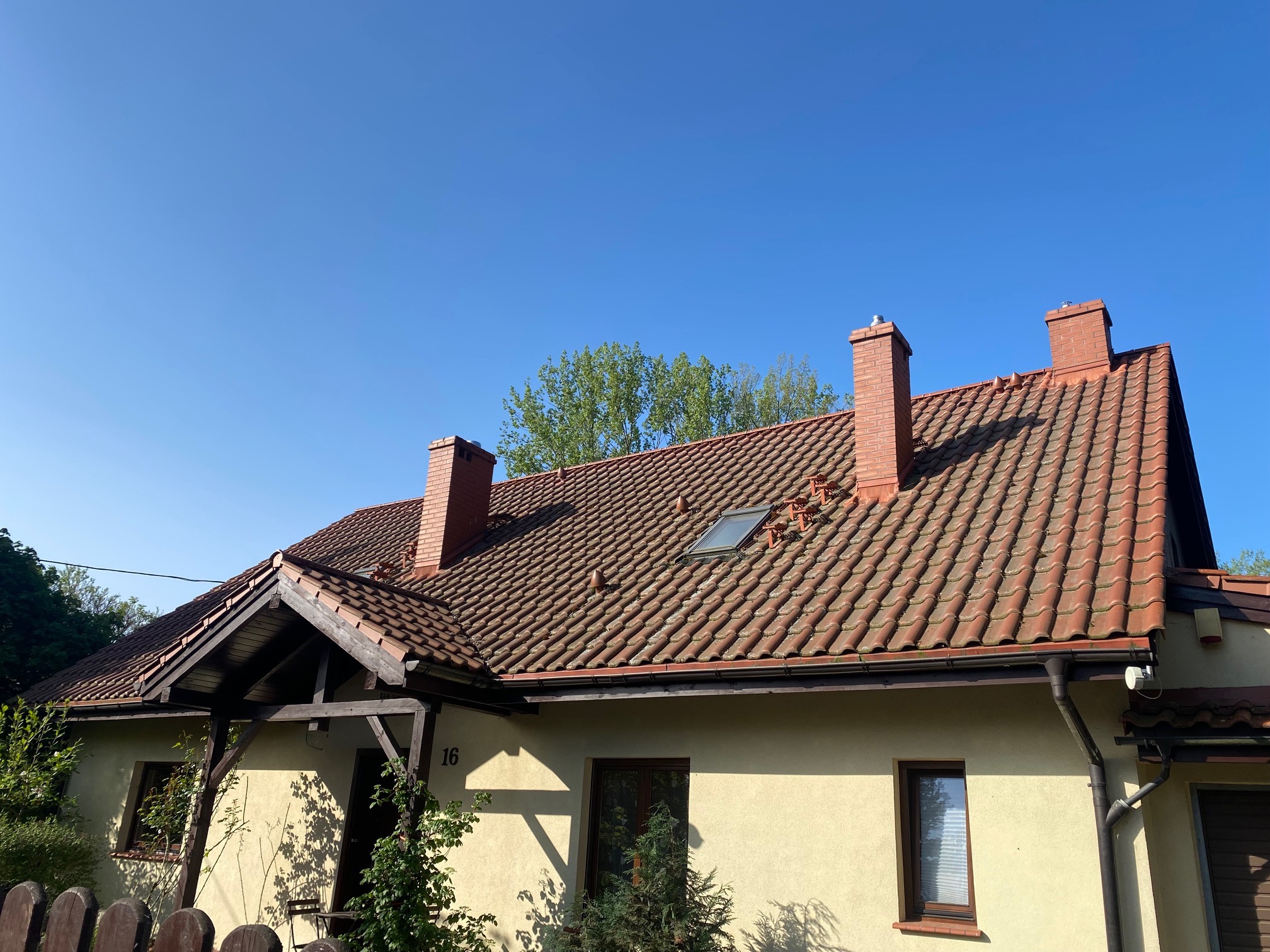 Dom jednorodzinny z czerwoną dachówką i czterema kominami, numer 16 na elewacji, błękitne niebo w tle. Widok z dołu, wczesne popołudnie.