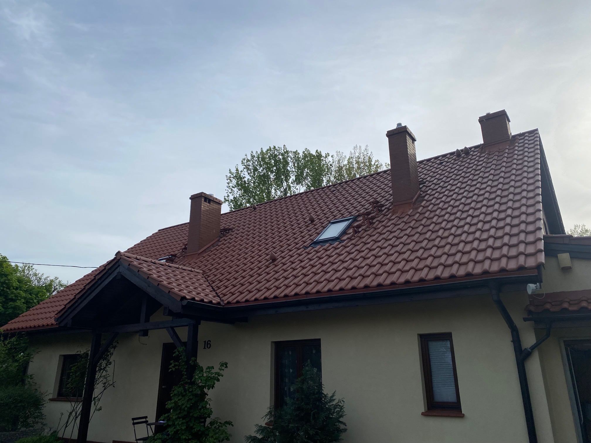 Dom jednorodzinny z nową, czerwoną dachówką i trzema kominami. Widoczny fragment elewacji w jasnym odcieniu. Numer domu '16' przy wejściu.