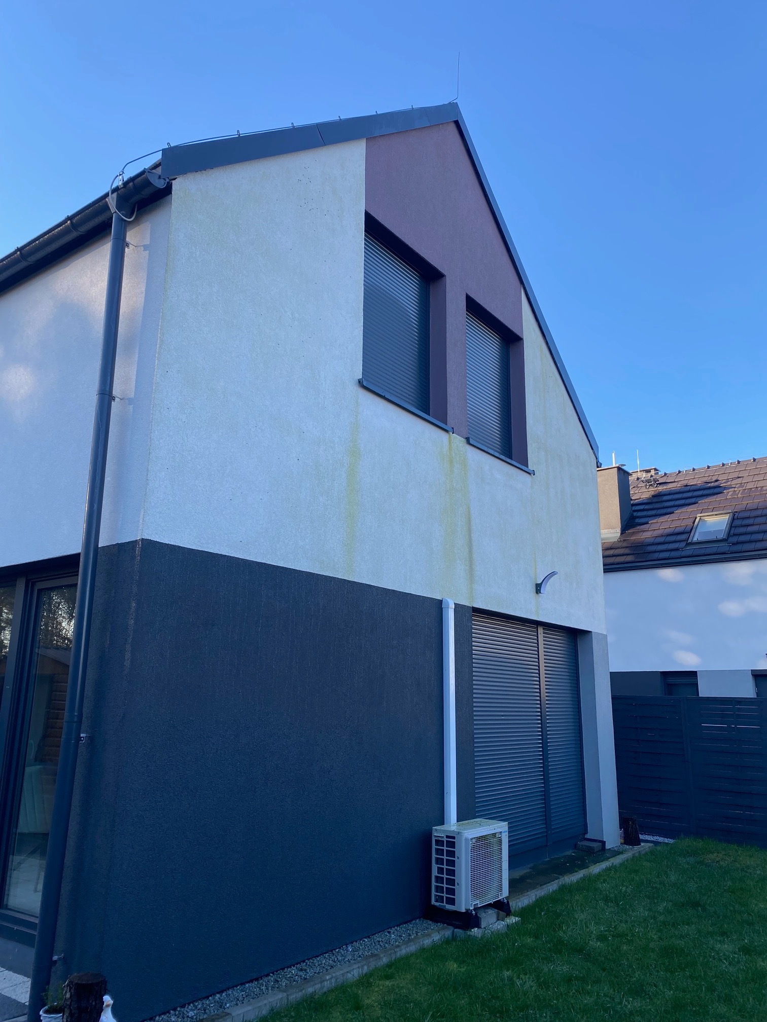 Dwukondygnacyjny dom z widocznymi zabrudzeniami na elewacji, z szarą dolną częścią i jasną górną, okna z roletami, trawnik i klimatyzator na zewnątrz.