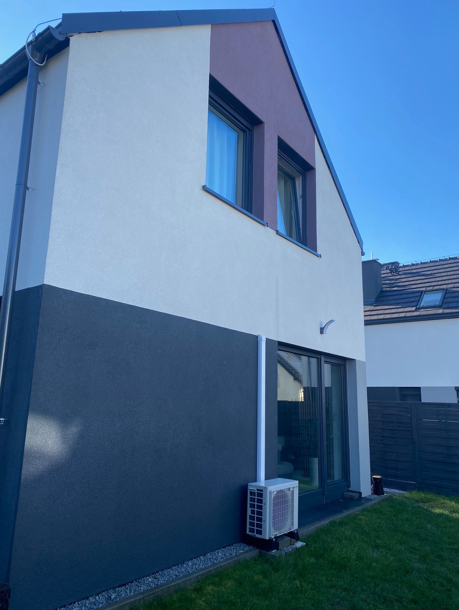 Dwukolorowa elewacja domu jednorodzinnego z klimatyzatorem na trawniku. Widoczne okna, drzwi tarasowe i fragment dachu sąsiedniego budynku. Błękitne niebo w tle.