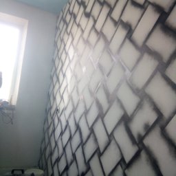 Aleksander Burkenia SPOKOBUD - Ściana z efektem cegieł w odcieniach szarości, wykonana techniką malarską. Widoczne narzędzia malarskie w tle. Nietypowy wzór na ścianie w pokoju.