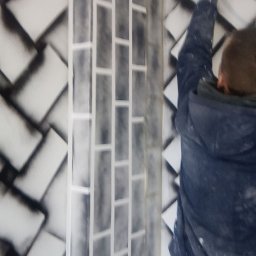 Aleksander Burkenia SPOKOBUD - Malowanie ściany z efektem cegieł przez malarza, widoczny fragment ściany z wzorem i osoba w trakcie pracy, trzymająca wałek malarski.