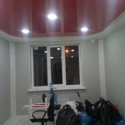 Aleksander Burkenia SPOKOBUD - Pokój po remoncie: czerwony, błyszczący sufit podwieszany z oświetleniem punktowym, szare ściany z białymi listwami i okno. Widoczne worki i narzędzia.