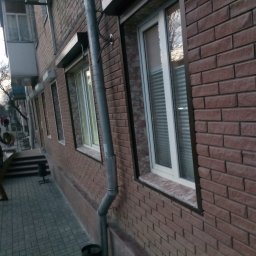 Aleksander Burkenia SPOKOBUD - Elewacja budynku z brązowej cegły z oknami PCV i rurą spustową. Na chodniku przechodnie. Ujęcie z dołu, lekko z boku, widoczne drzewa w tle.