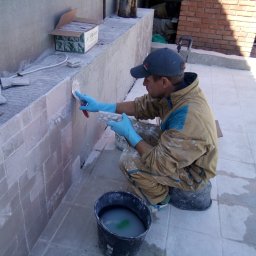 Aleksander Burkenia SPOKOBUD - Mężczyzna w niebieskich rękawicach nakłada zaprawę na beżowe płytki na ścianie, obok wiadro i karton z płytkami. Praca na zewnątrz w słoneczny dzień.