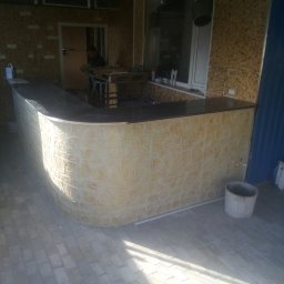 Aleksander Burkenia SPOKOBUD - Wykończony bar z dekoracyjną, teksturowaną ścianą imitującą kamień, z ciemnym blatem, na tle pomieszczenia z oknem i częściowo wykończonymi ścianami.
