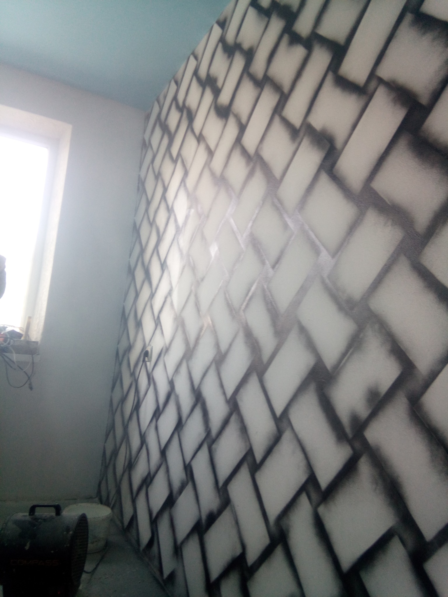 Ściana z efektem cegieł w odcieniach szarości, wykonana techniką malarską. Widoczne narzędzia malarskie w tle. Nietypowy wzór na ścianie w pokoju.