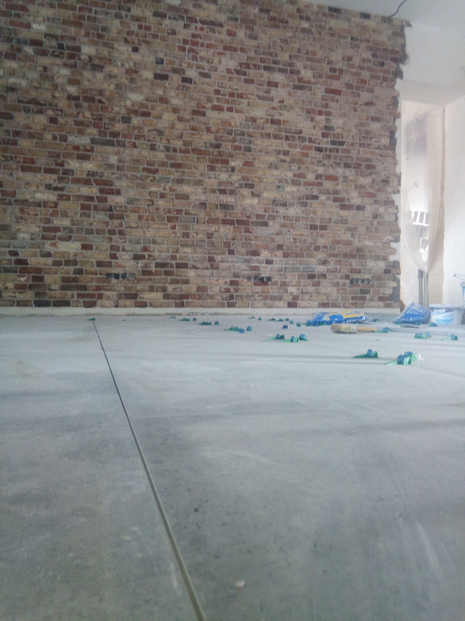 Surowa ściana z cegły i szara podłoga w trakcie prac wykończeniowych. Widoczne narzędzia i akcesoria glazurnicze na podłodze. W tle jasne pomieszczenie.