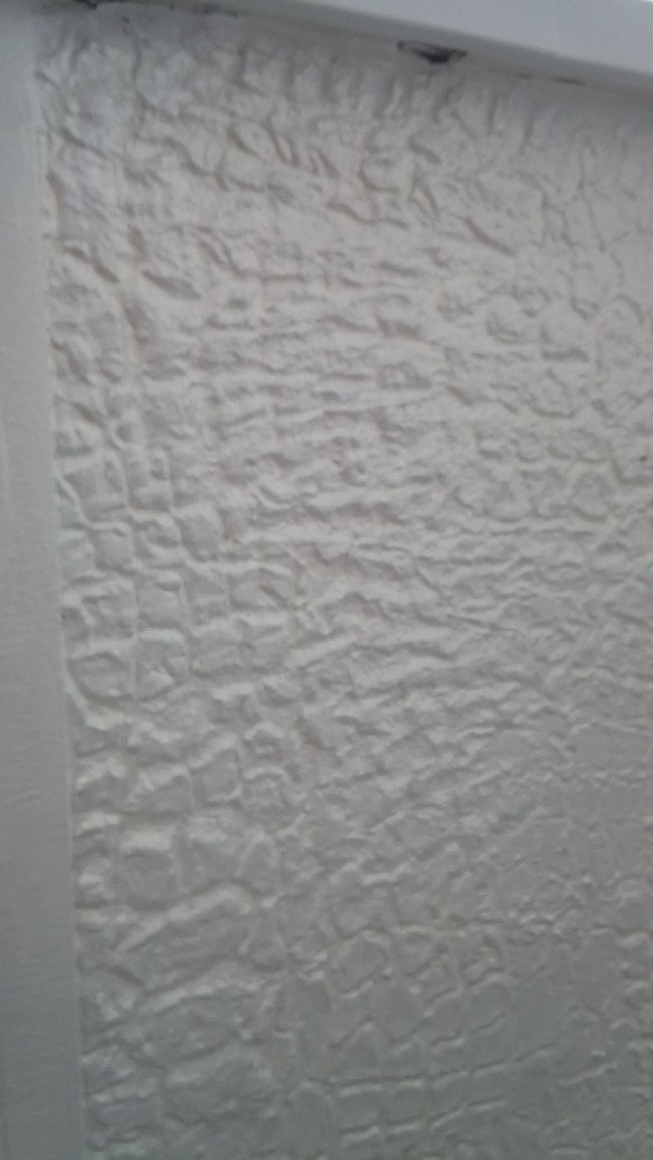 Biała ściana z wyraźną, nieregularną fakturą przypominającą kamień, oświetlona z góry, z fragmentem białej ramy po lewej stronie kadru.