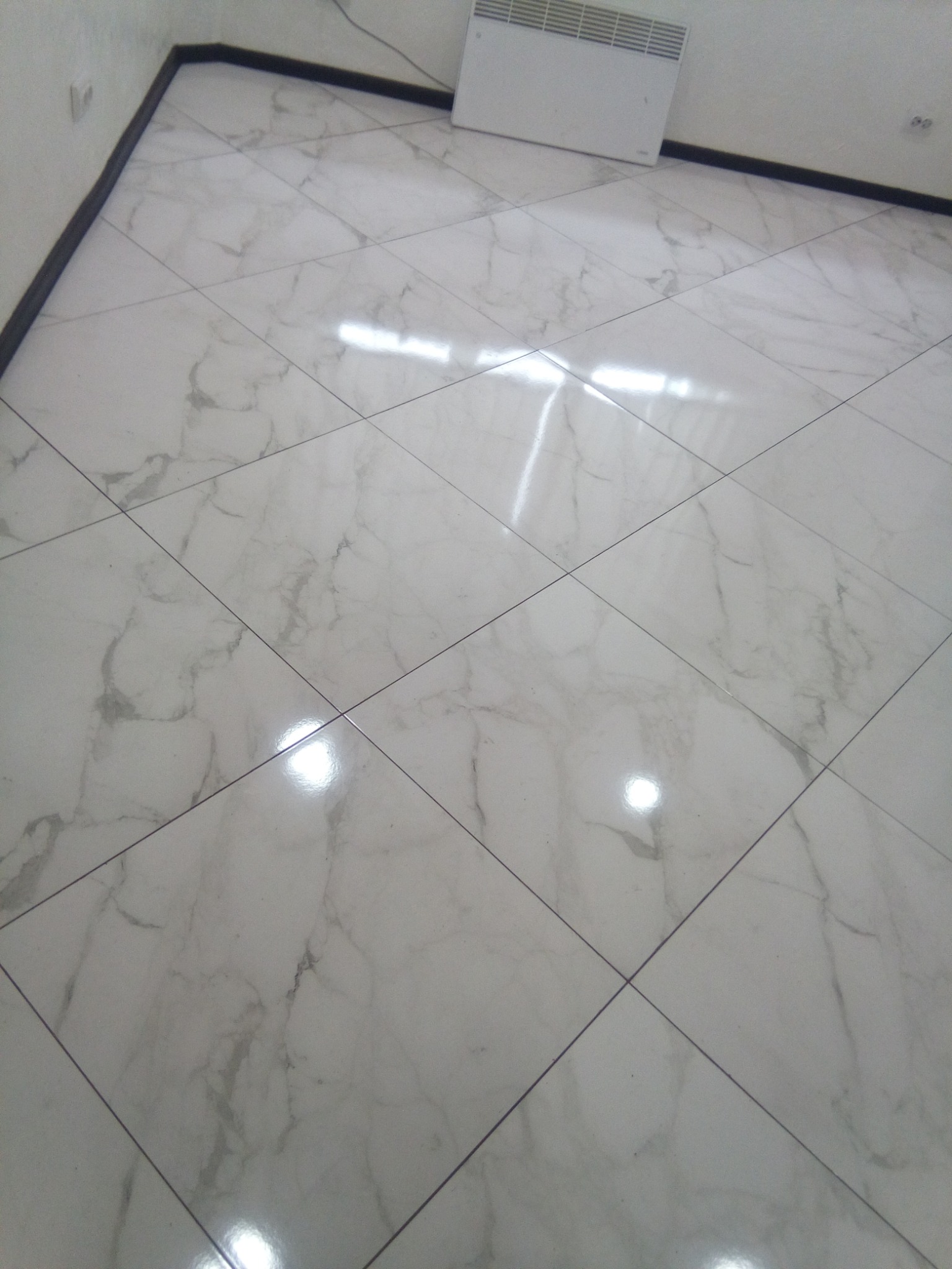 Błyszcząca, biała podłoga z płytek imitujących marmur, ułożonych w kwadraty. Widoczny fragment grzejnika na ścianie i ciemna listwa przypodłogowa.