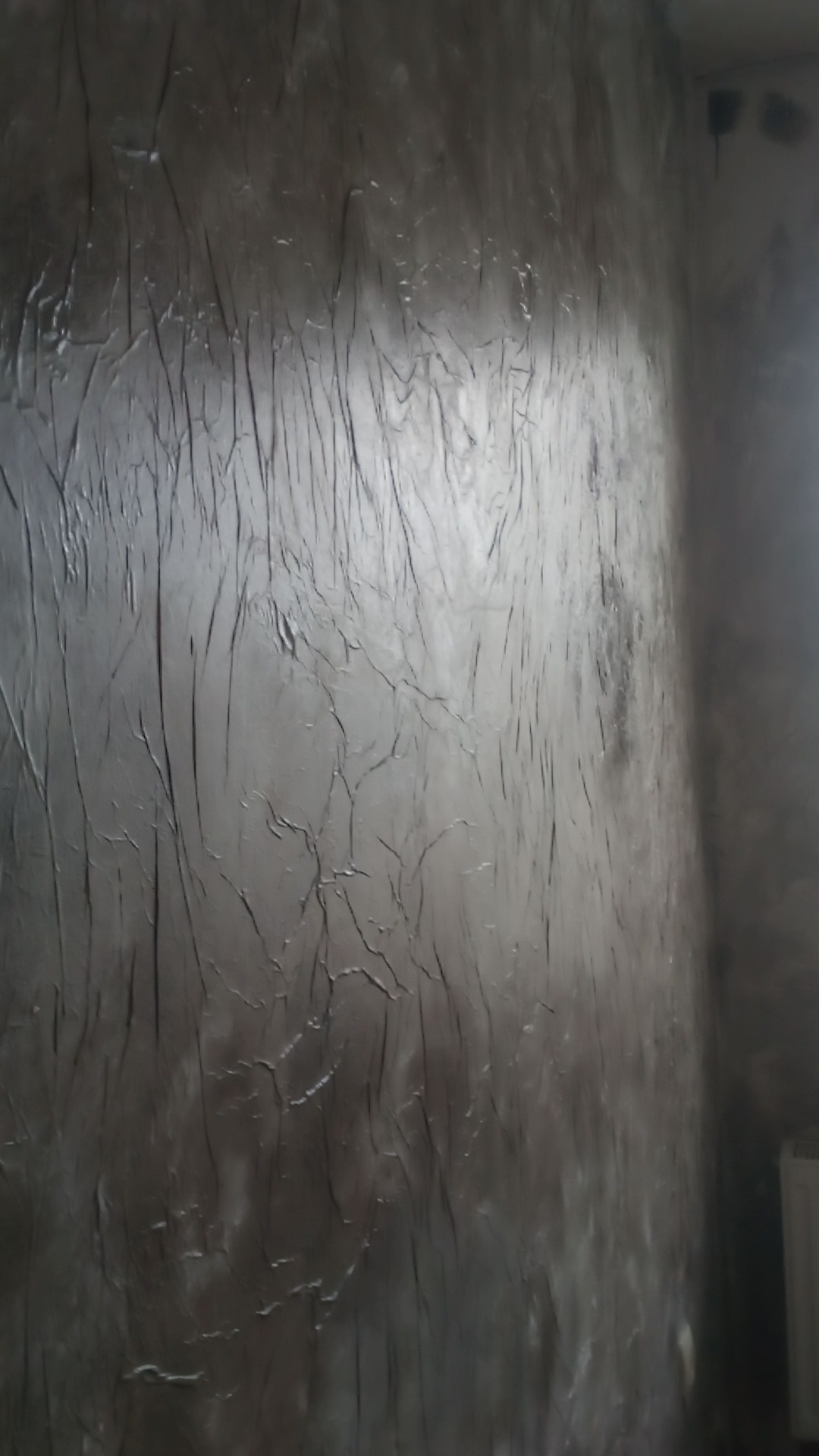 Ściana z dekoracyjną, szarą strukturą przypominającą popękany beton. Widoczne pionowe smugi i nieregularne pęknięcia. Oświetlenie boczne podkreśla fakturę.