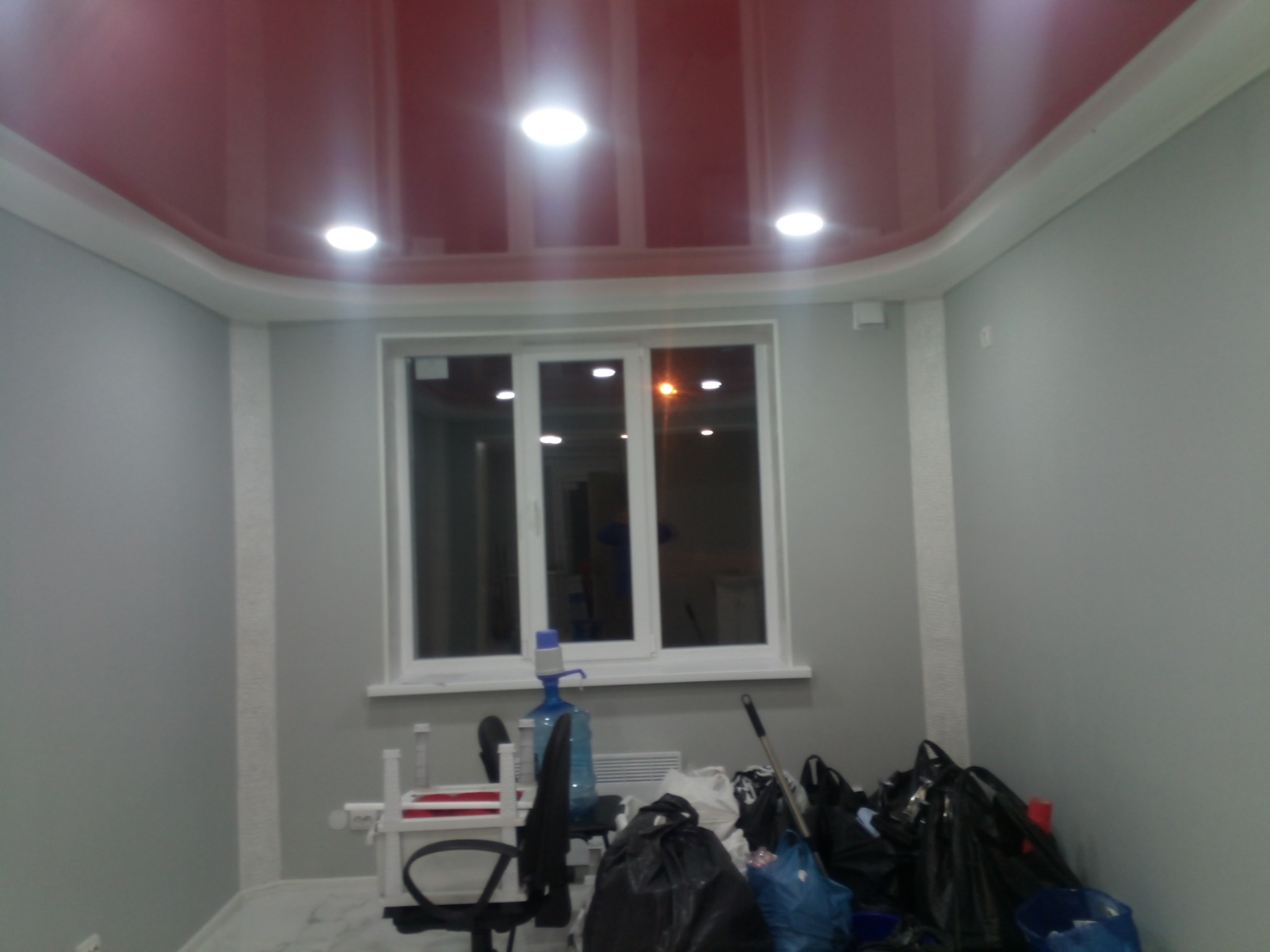 Pokój po remoncie: czerwony, błyszczący sufit podwieszany z oświetleniem punktowym, szare ściany z białymi listwami i okno. Widoczne worki i narzędzia.
