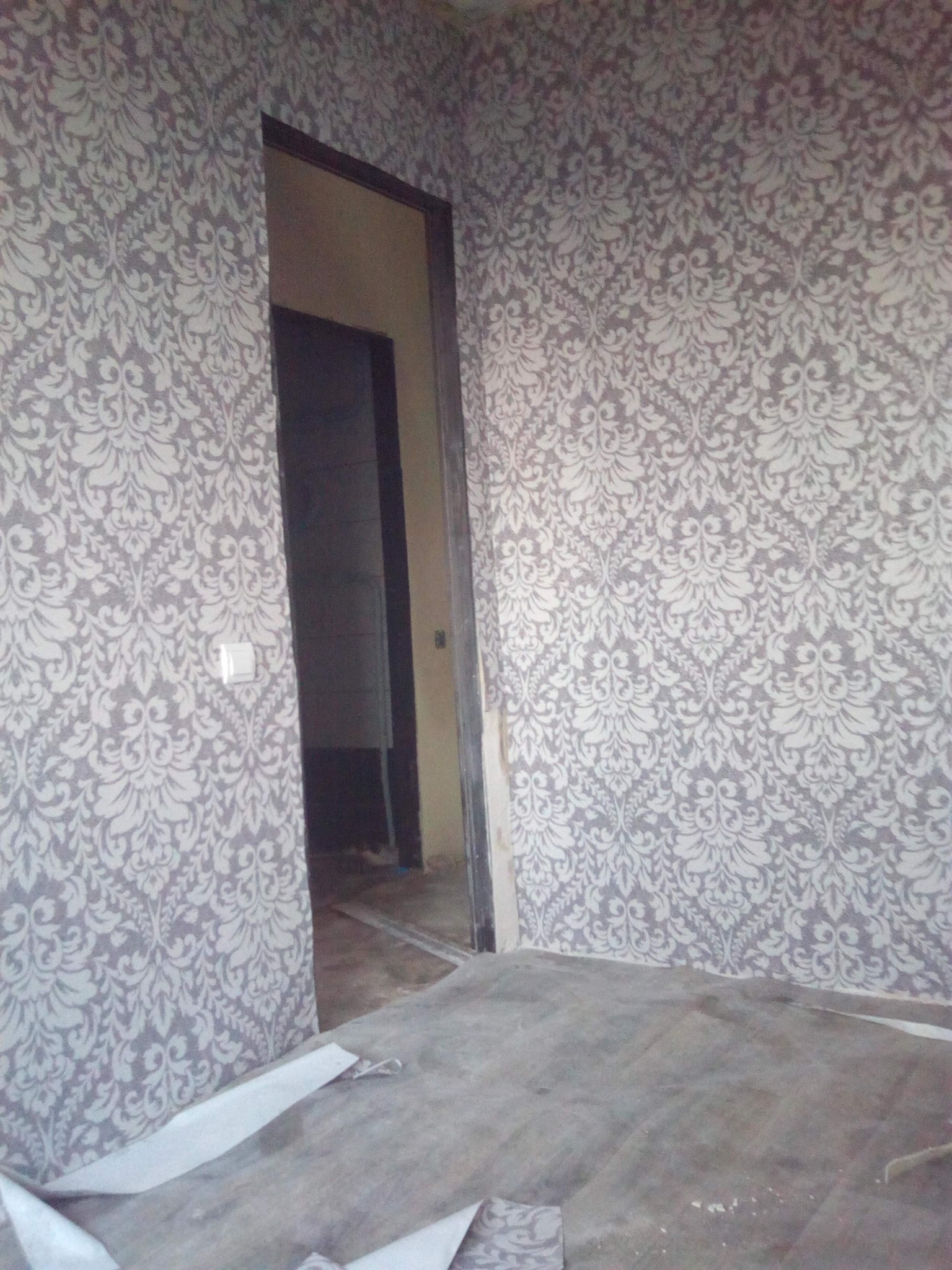 Ściana z wzorzystą tapetą w odcieniach szarości i bieli, widoczny fragment podłogi i otwór drzwiowy z ciemną ościeżnicą. Fragment tapety leży na podłodze.