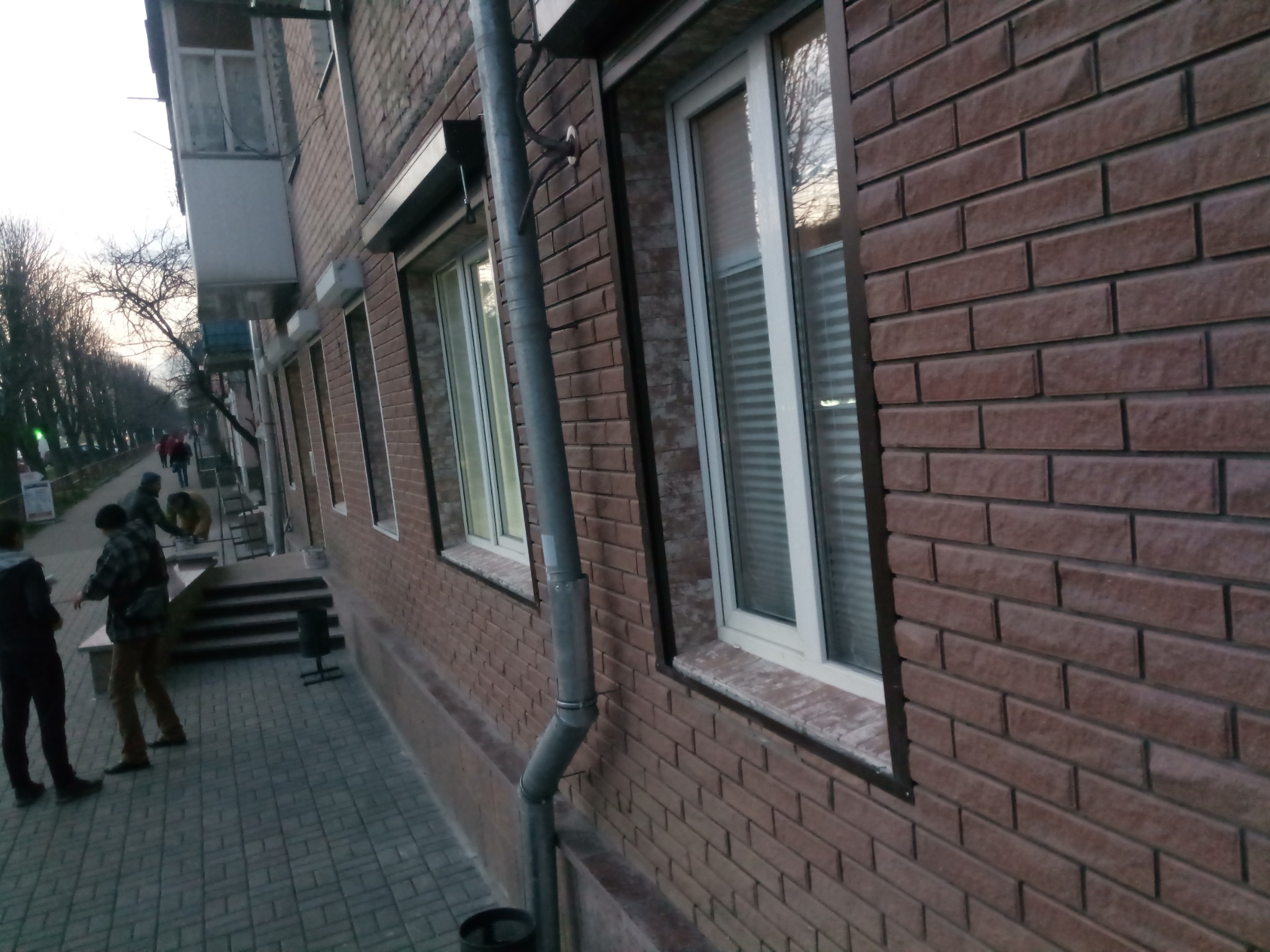 Elewacja budynku z brązowej cegły z oknami PCV i rurą spustową. Na chodniku przechodnie. Ujęcie z dołu, lekko z boku, widoczne drzewa w tle.