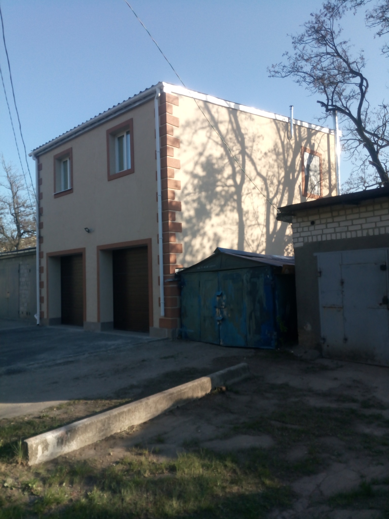 Dwukondygnacyjny budynek z beżową elewacją i brązowymi detalami okien i narożników, sąsiadujący z garażami, w słoneczny dzień.