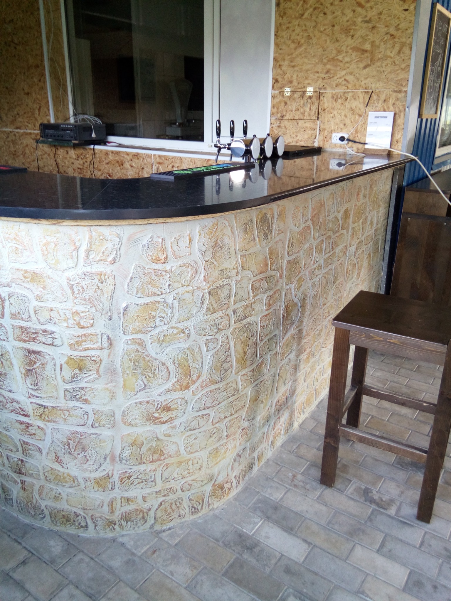 Bar z dekoracyjną ścianą imitującą kamień i ciemnym blatem, wysoki drewniany stołek. Podłoga wyłożona płytkami imitującymi cegłę. Widoczne krany do piwa.