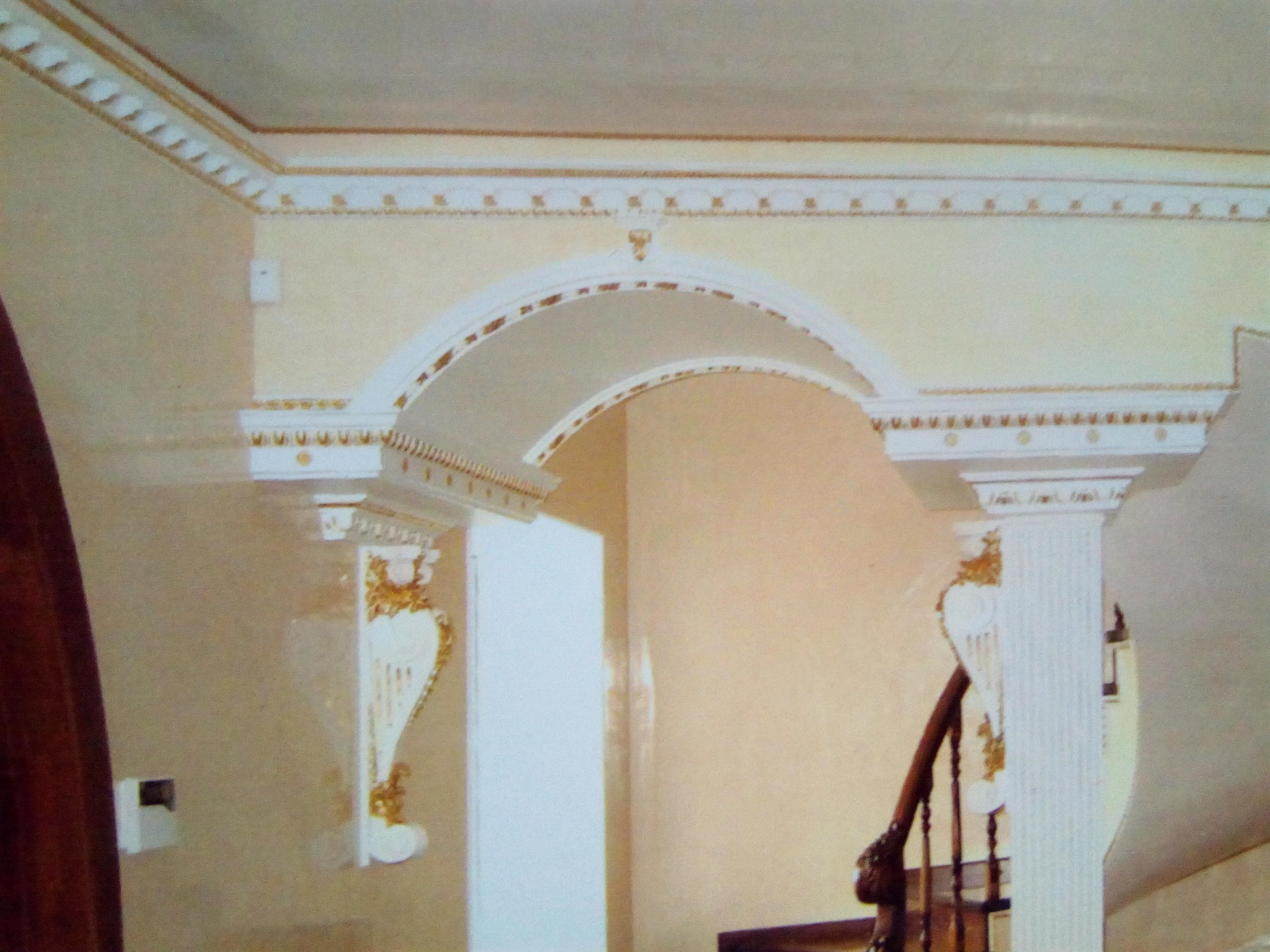 Eleganckie wykończenie wnętrza z dekoracyjnym łukiem i kolumnami w odcieniach bieli i złota, widoczne detale sztukaterii i fragment schodów.