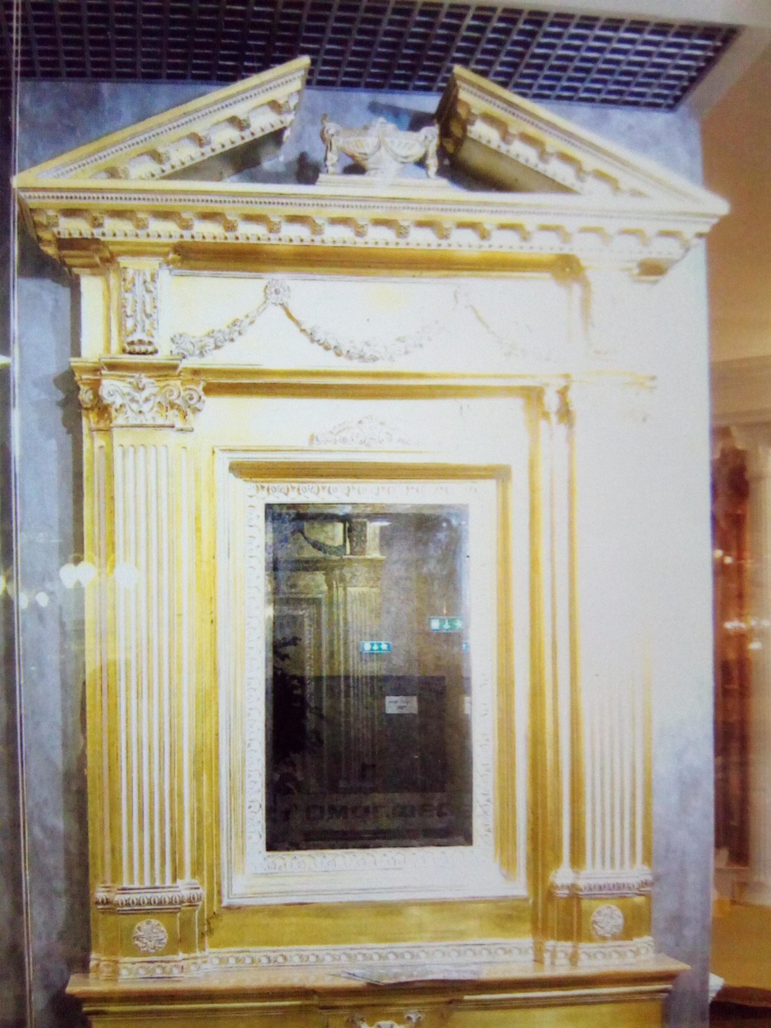 Złota, bogato zdobiona rama lustra w stylu klasycystycznym, z kolumnami i ornamentami, odbijająca wnętrze pomieszczenia.