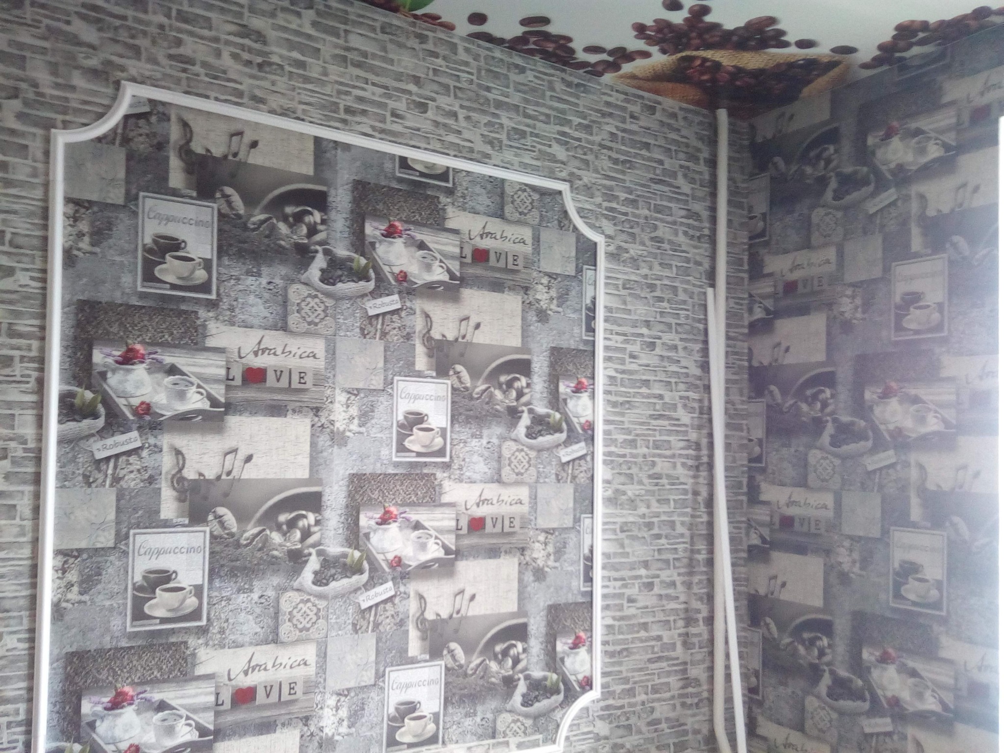 Ściana z tapetą w motywy kawowe, obramowana białą sztukaterią. Na suficie dekoracja z ziaren kawy. Widoczny fragment rury. Nietypowy wzór i aranżacja.