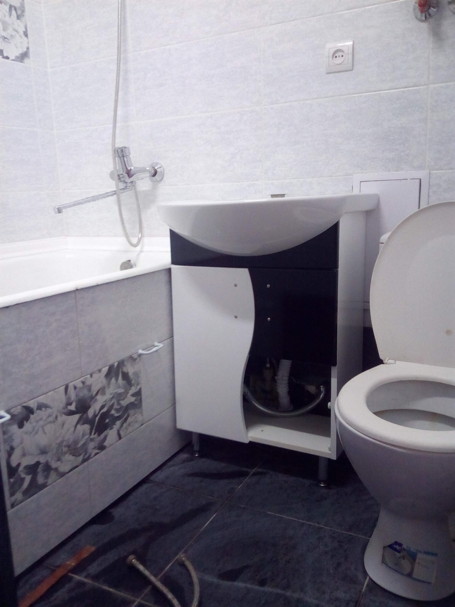 Umywalka z szafką w łazience z biało-czarnymi frontami, obok toaleta i fragment wanny z szarą mozaiką. Widoczne płytki na ścianach i ciemna podłoga.