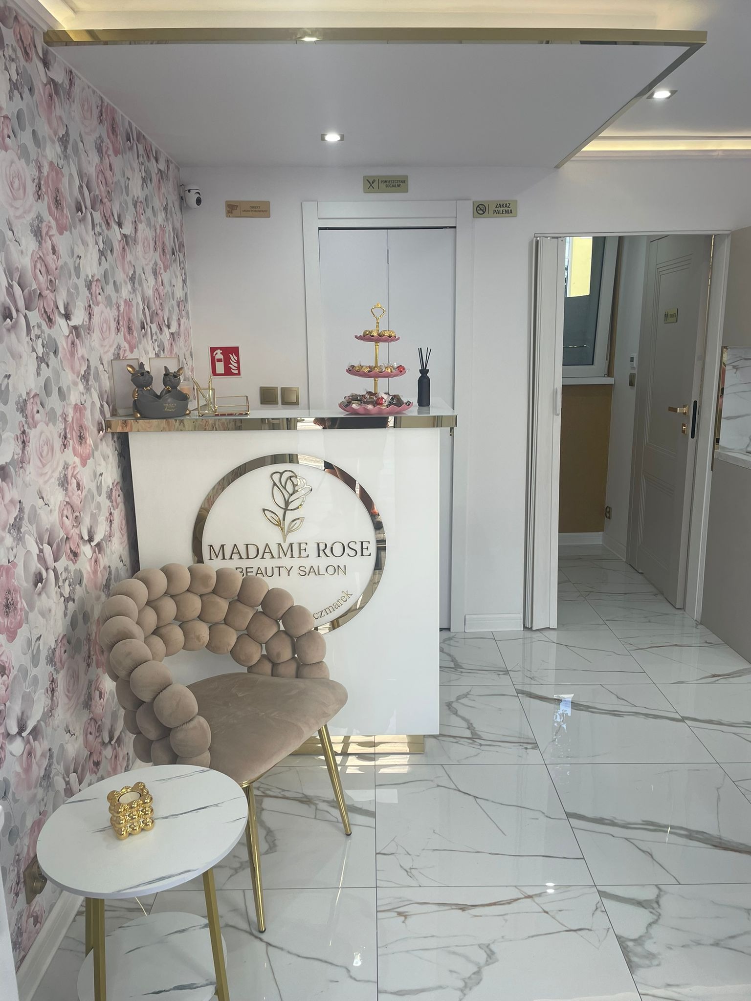 Eleganckie wnętrze salonu kosmetycznego Madame Rose z recepcją, krzesłem w stylu glamour i marmurową podłogą. W tle ściana w kwiaty i dekoracje.