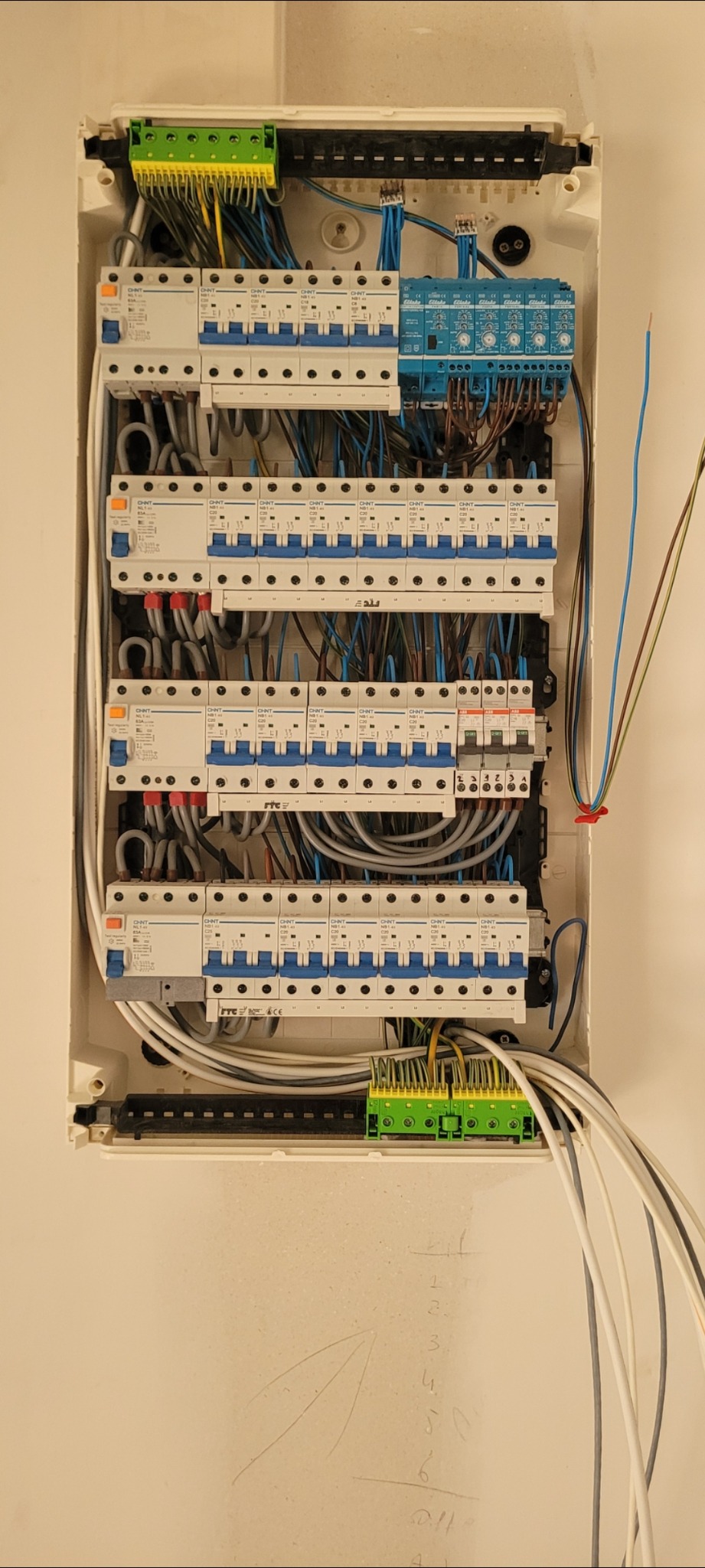 Rozdzielnia elektryczna w białej obudowie z widocznymi bezpiecznikami, przewodami i listwami zaciskowymi. Widoczne ślady ołówka na ścianie.