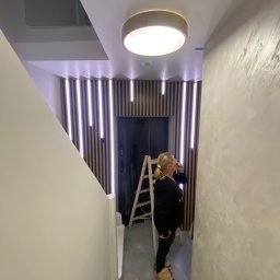 Przedsiębiorstwo wielobranżowe Emilia Senger - Nowoczesne wykończenie klatki schodowej z drewnianymi lamelami i oświetleniem LED. Kobieta z psem przy drabinie. Ściana z efektem dekoracyjnym.