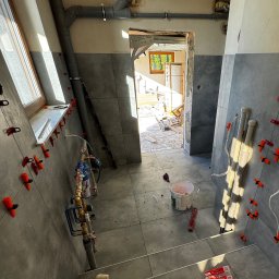 Przedsiębiorstwo wielobranżowe Emilia Senger - Szare płytki na ścianach i podłodze w trakcie montażu. Widoczne narzędzia glazurnicze, rury i okno. Prace wykończeniowe w łazience w toku.