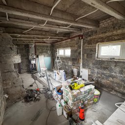 Przedsiębiorstwo wielobranżowe Emilia Senger - Wnętrze w trakcie remontu: surowe ściany z cegły, materiały budowlane, narzędzia, instalacje. Widoczne okna i elementy instalacji grzewczej. Etap prac wykończeniowych.