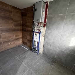 Przedsiębiorstwo wielobranżowe Emilia Senger - Szara i brązowa glazura w łazience, z widoczną instalacją podtynkową WC i piecem gazowym. Ułożone płytki podłogowe i ścienne. Etap prac wykończeniowych.