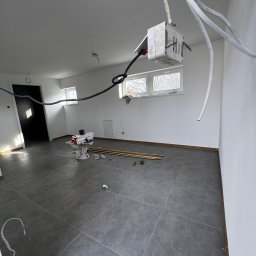 Przedsiębiorstwo wielobranżowe Emilia Senger - Wnętrze w trakcie prac: szare płytki podłogowe, białe ściany, wystające kable elektryczne, rury i narzędzia. Widok na okno i drzwi w głębi pomieszczenia.