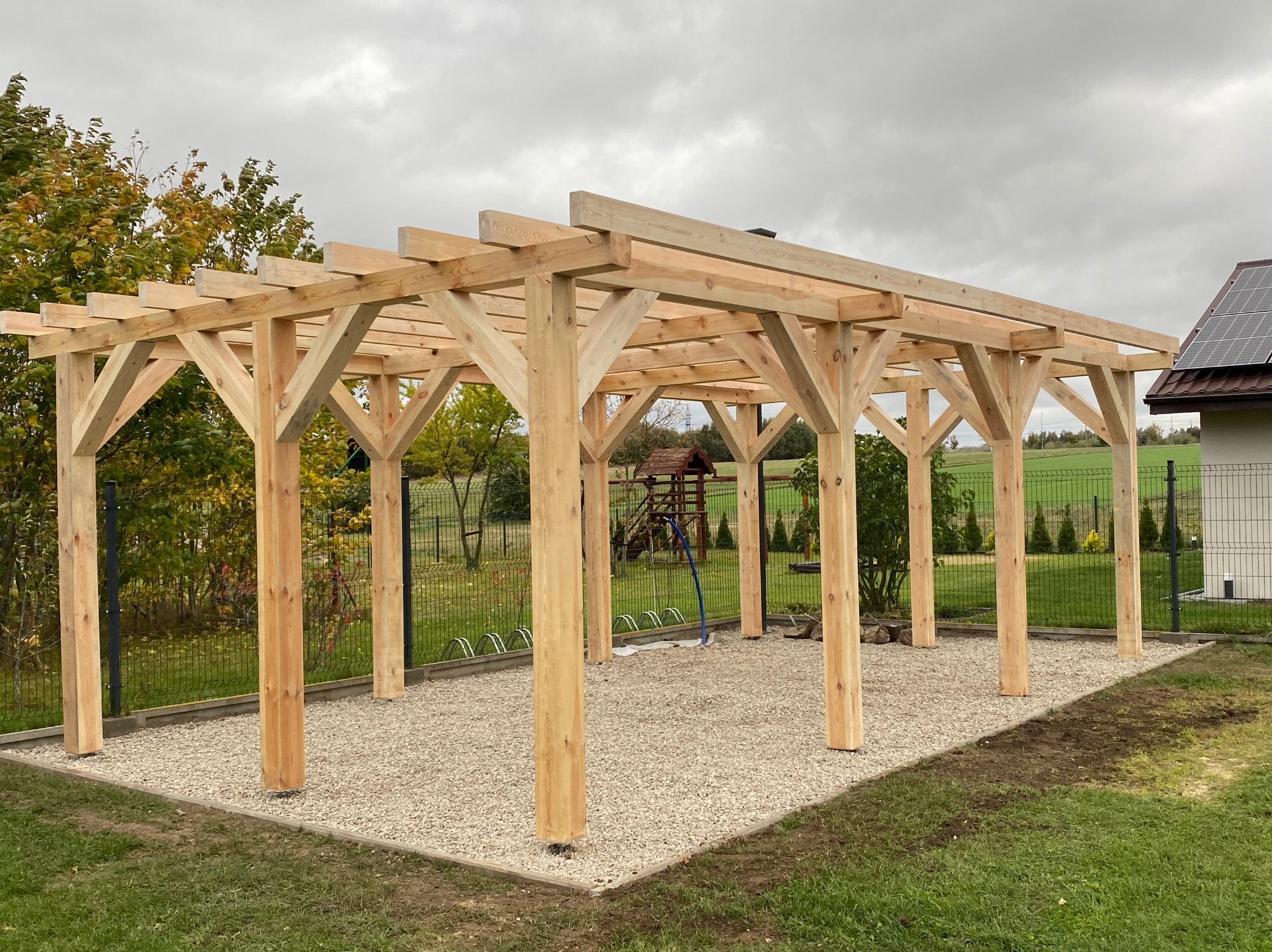 Drewniana pergola z jasnego drewna na żwirowym podłożu, widok z zewnątrz, w tle zieleń i dom z panelami słonecznymi. Konstrukcja w surowym stanie, bez zadaszenia.