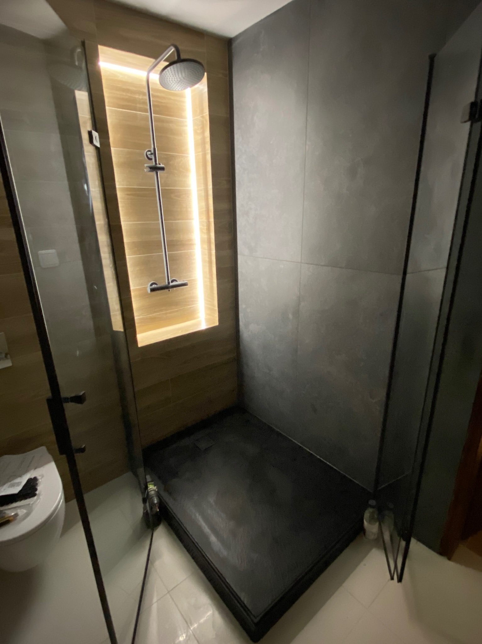 Nowoczesna kabina prysznicowa walk-in z czarnym brodzikiem i szarą ścianą, oświetlona wnęka z drewnopodobnymi płytkami. Widoczna toaleta i akcesoria.