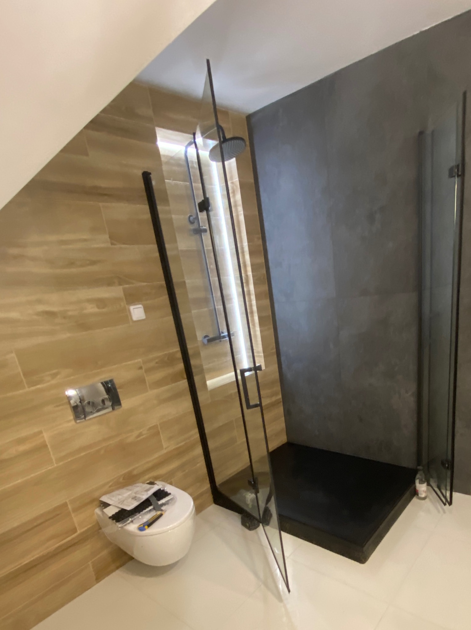 Nowoczesna łazienka po remoncie: kabina prysznicowa typu walk-in z czarnymi profilami, drewnopodobne płytki na ścianie i szara ściana. Umywalka podwieszana.