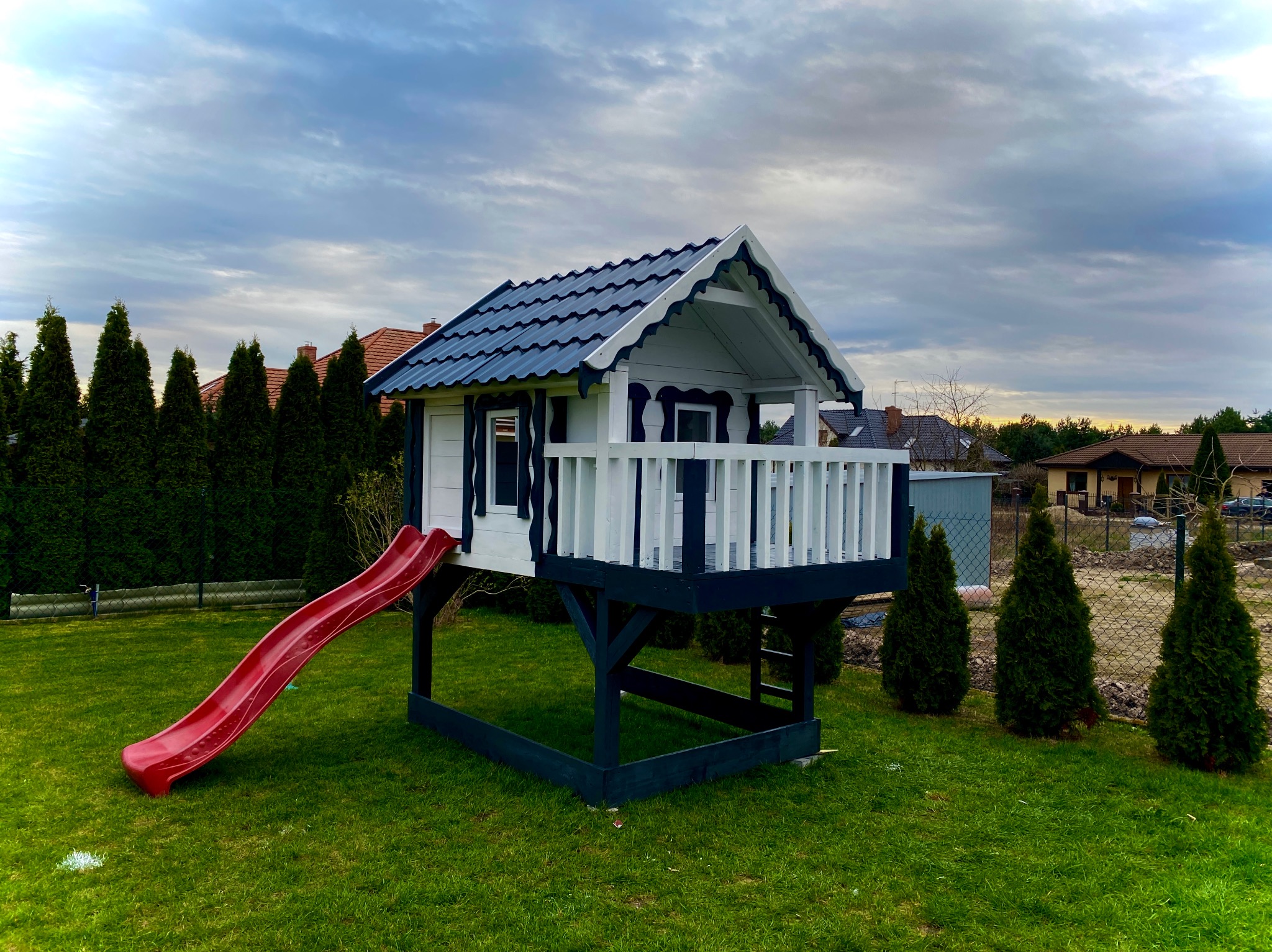 Drewniany domek dla dzieci z czerwoną zjeżdżalnią, na podwyższeniu, z niebieskim dachem, otoczony zielonym trawnikiem i tujami, w tle zabudowa jednorodzinna.