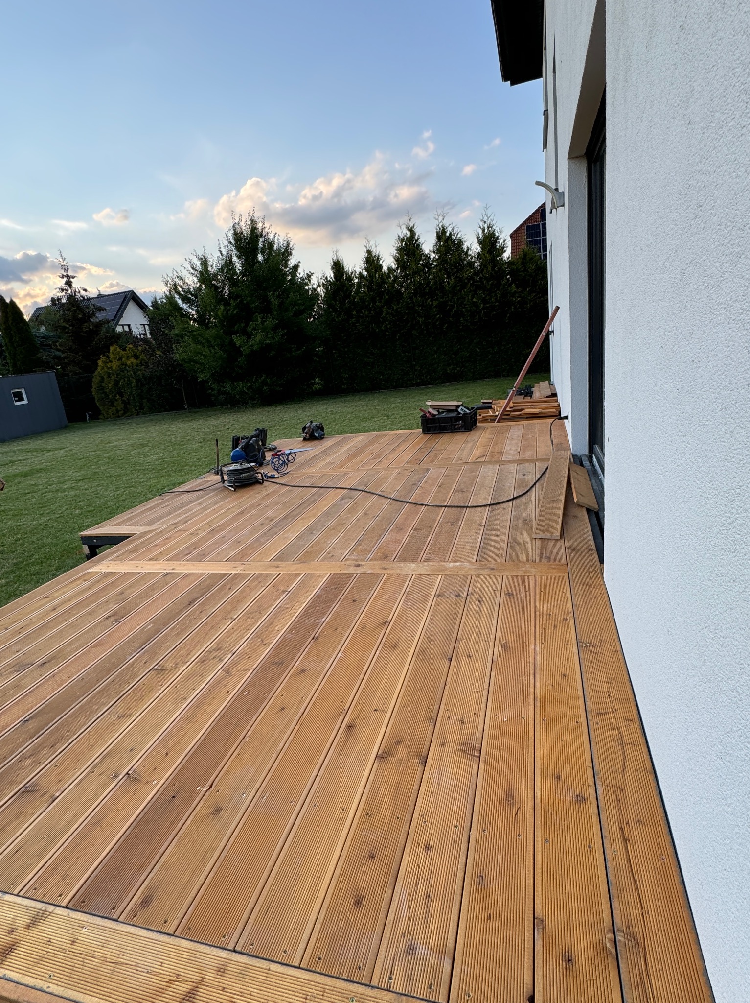Drewniany taras w trakcie budowy, deski ułożone, widoczne narzędzia i materiały, dom z białym tynkiem w tle, zielony trawnik i drzewa. Ujęcie z boku, wczesny wieczór.