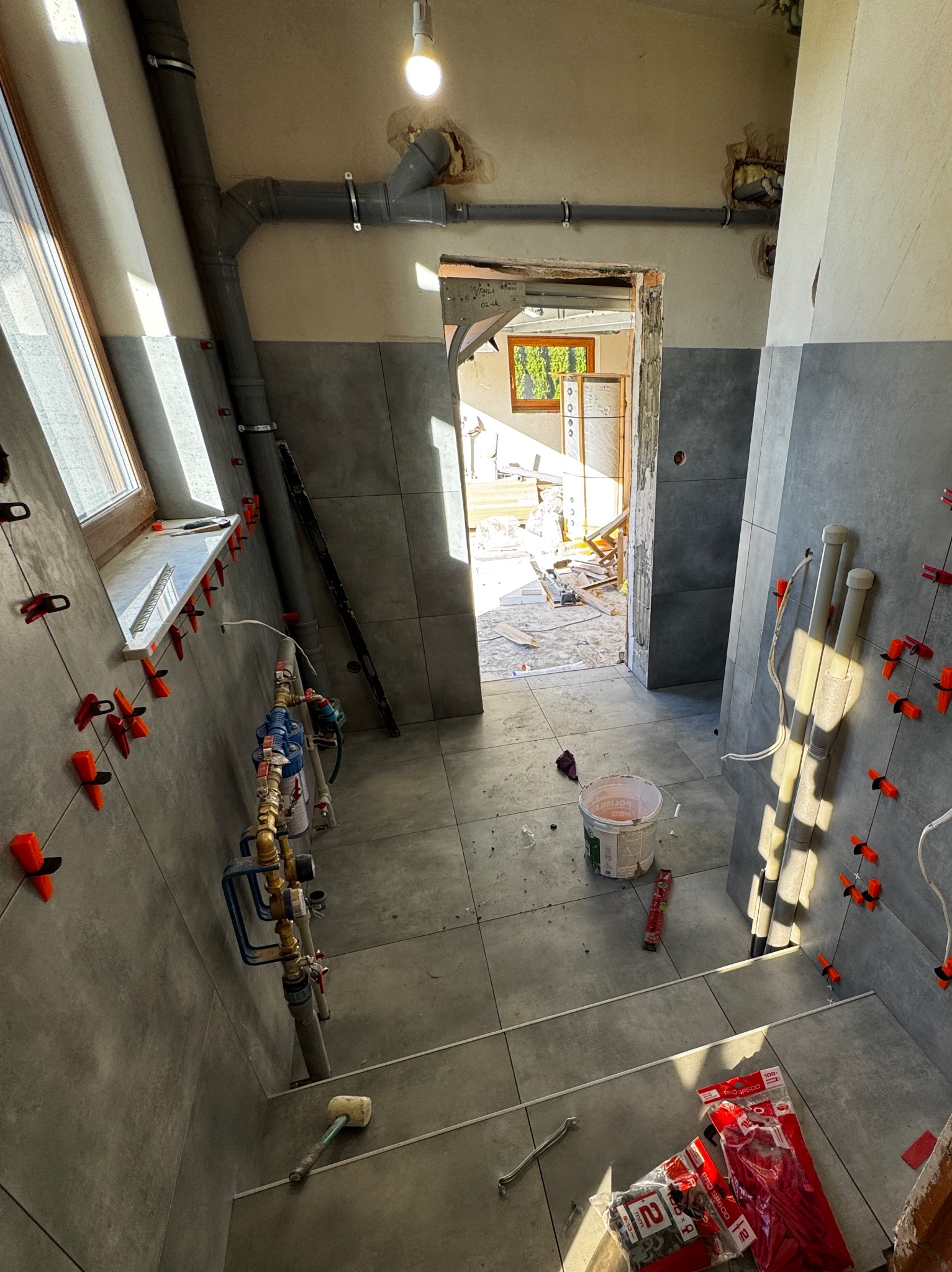 Szare płytki na ścianach i podłodze w trakcie montażu. Widoczne narzędzia glazurnicze, rury i okno. Prace wykończeniowe w łazience w toku.