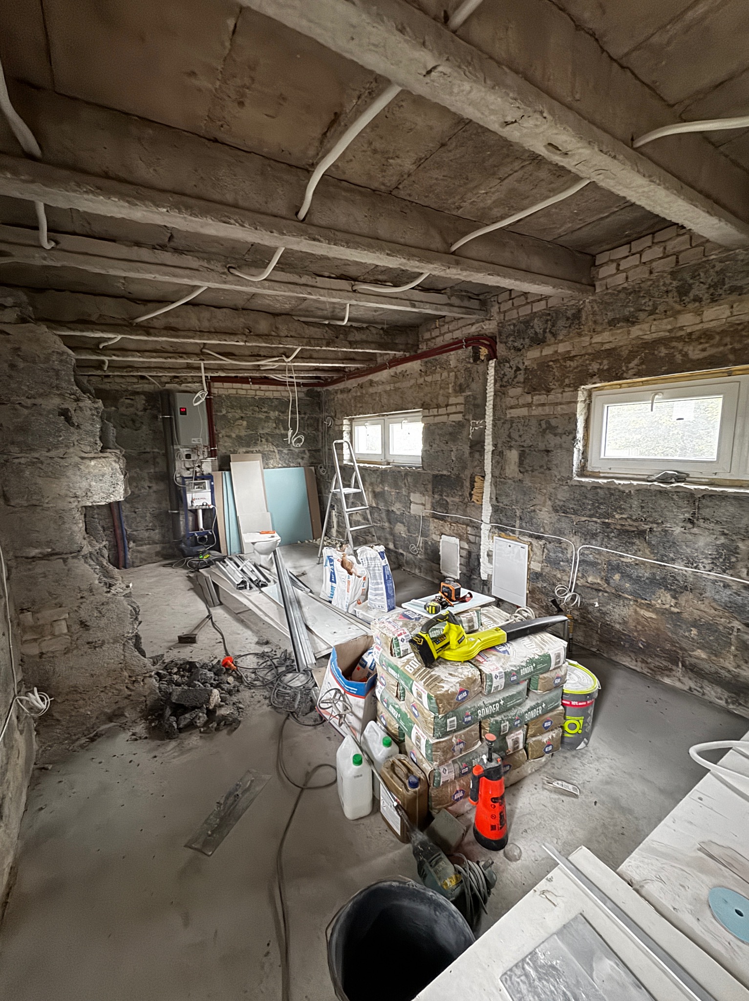 Wnętrze w trakcie remontu: surowe ściany z cegły, materiały budowlane, narzędzia, instalacje. Widoczne okna i elementy instalacji grzewczej. Etap prac wykończeniowych.