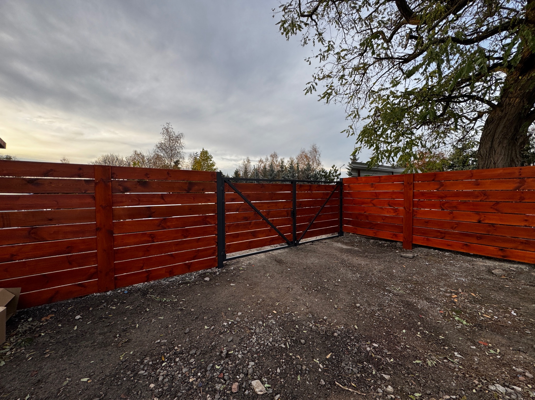 Nowoczesne ogrodzenie z drewnianych desek w kolorze ciepłego brązu, z czarną metalową bramą, na tle pochmurnego nieba i drzew z jesiennymi liśćmi.