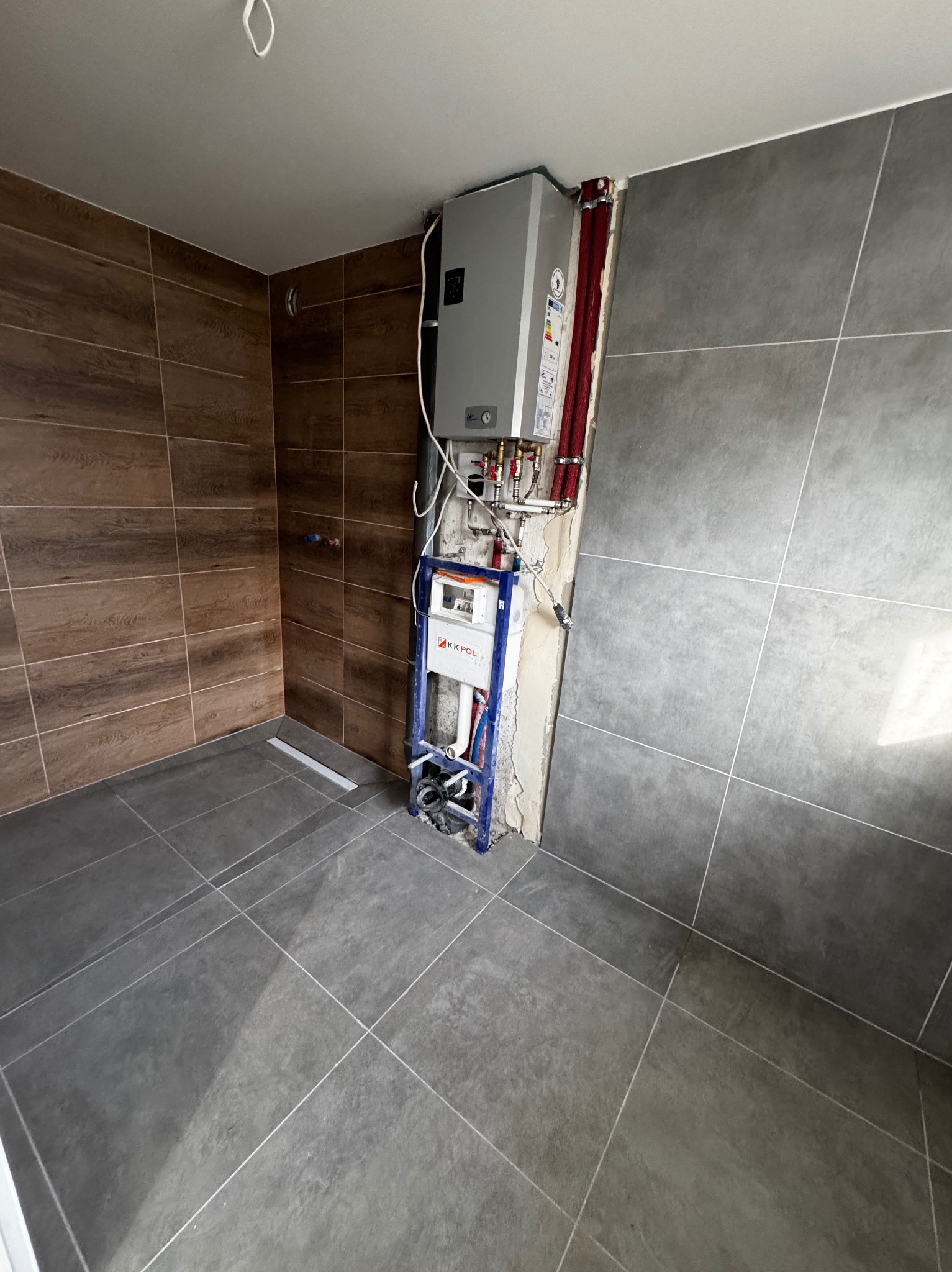 Szara i brązowa glazura w łazience, z widoczną instalacją podtynkową WC i piecem gazowym. Ułożone płytki podłogowe i ścienne. Etap prac wykończeniowych.