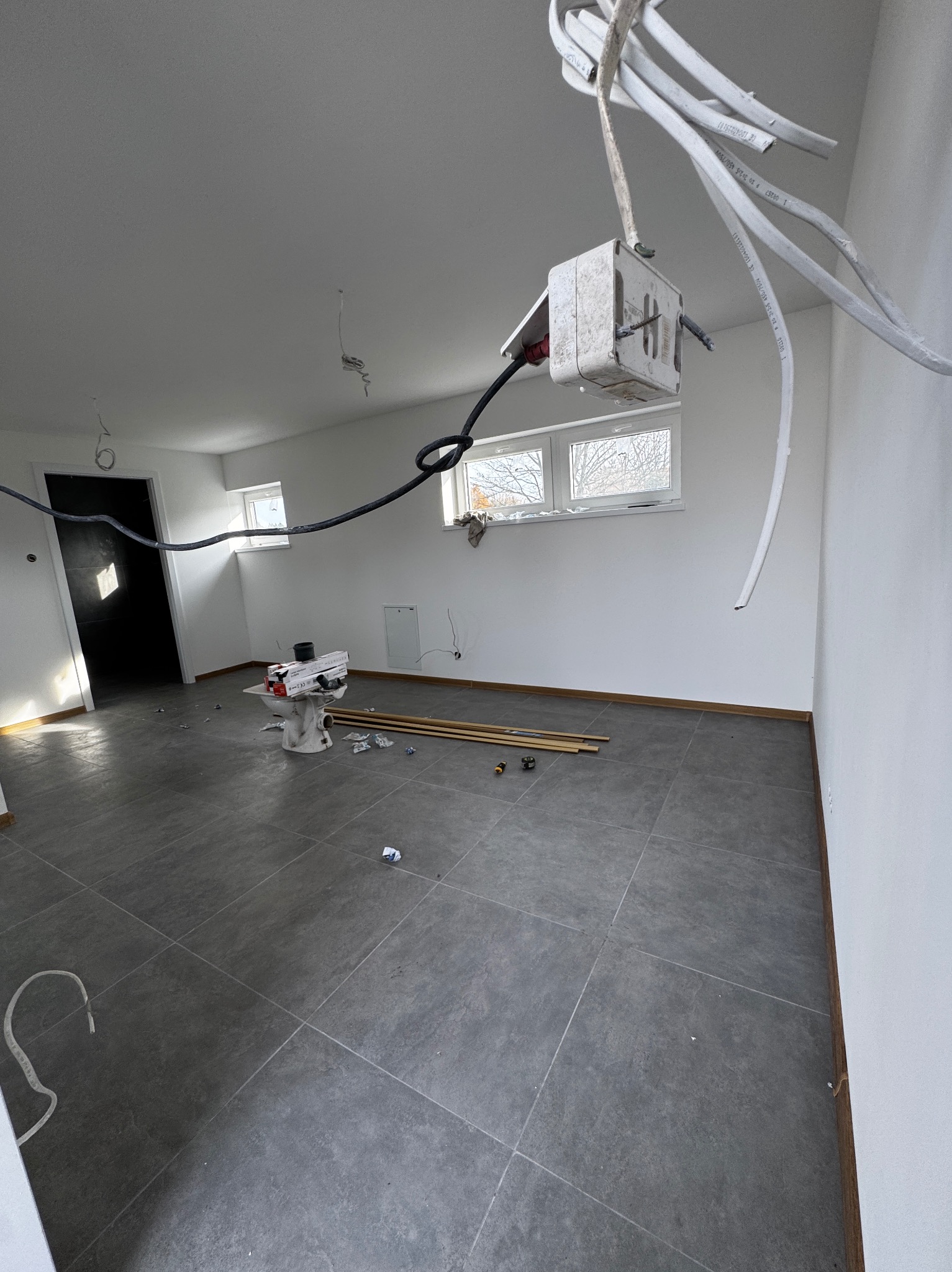 Wnętrze w trakcie prac: szare płytki podłogowe, białe ściany, wystające kable elektryczne, rury i narzędzia. Widok na okno i drzwi w głębi pomieszczenia.