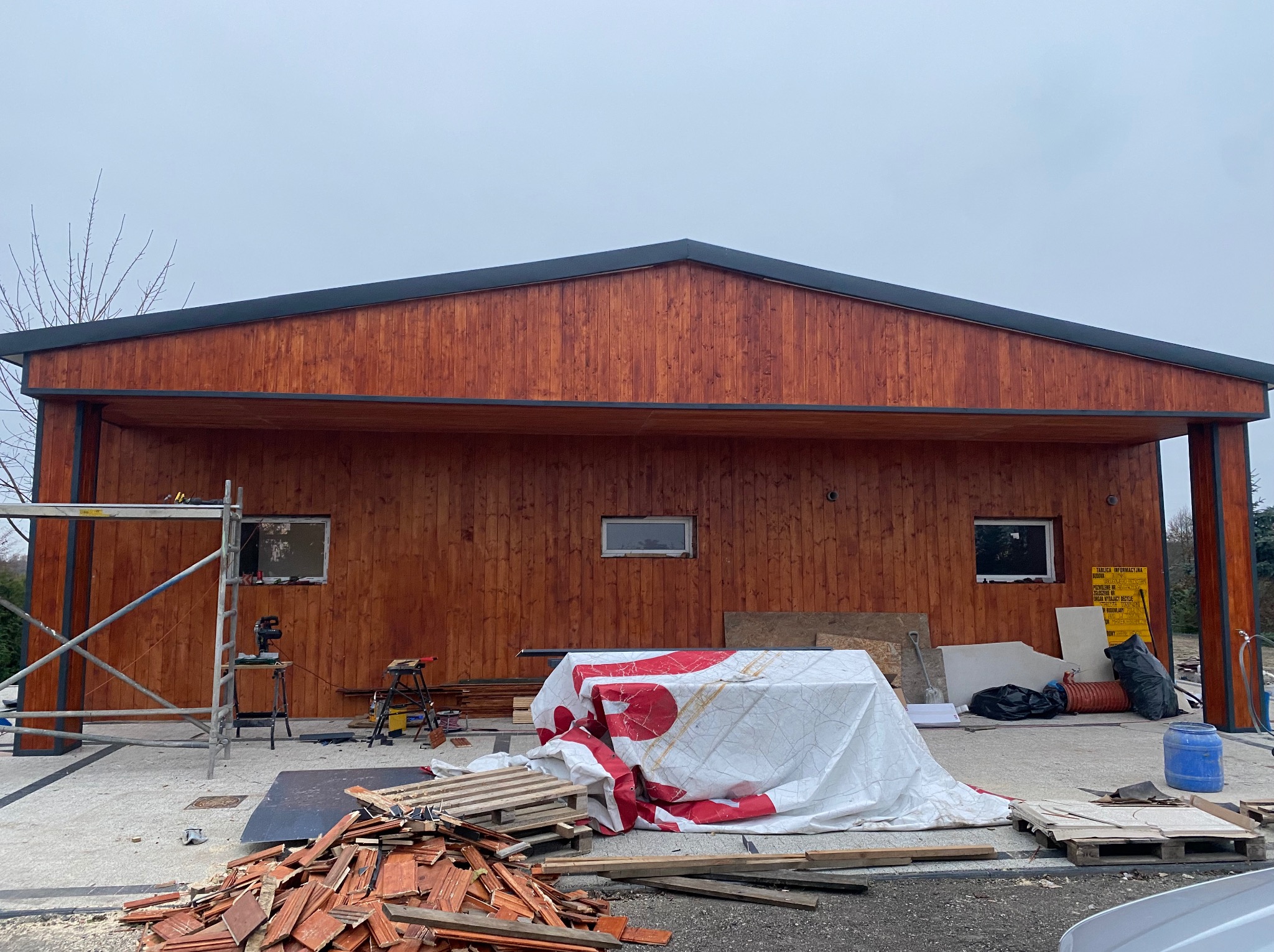 Drewniana konstrukcja z widocznymi śladami prac budowlanych, rusztowaniem i stertą dachówek. Widoczne okna i elementy wykończeniowe.