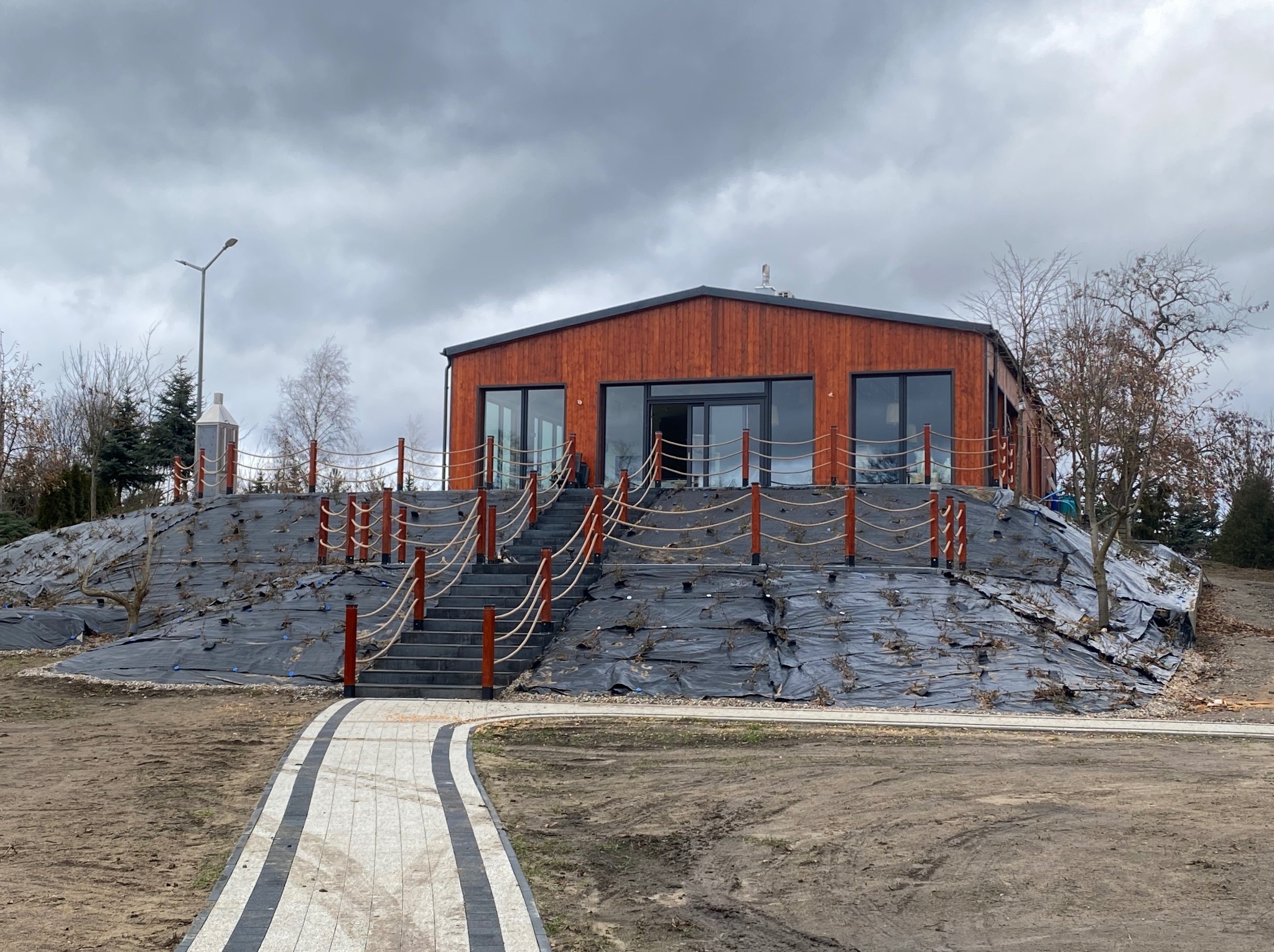 Nowoczesny dom z drewnianą elewacją, schodami i ścieżką z kostki brukowej na wzgórzu, zabezpieczonym czarną agrowłókniną. Pochmurne niebo w tle.