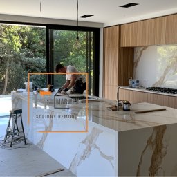 Marwex Remonty - Montaż blatu kuchennego z marmurowym wzorem przez pracownika w nowoczesnej kuchni z dużym oknem i drewnianymi szafkami. Widoczne narzędzia i drabina.