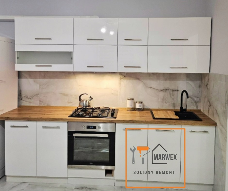 Biała, nowoczesna kuchnia z drewnianym blatem i marmurową ścianą. Widoczny piekarnik i płyta gazowa. Uwagę zwraca logo firmy na szafce.