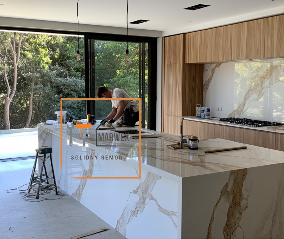 Montaż blatu kuchennego z marmurowym wzorem przez pracownika w nowoczesnej kuchni z dużym oknem i drewnianymi szafkami. Widoczne narzędzia i drabina.