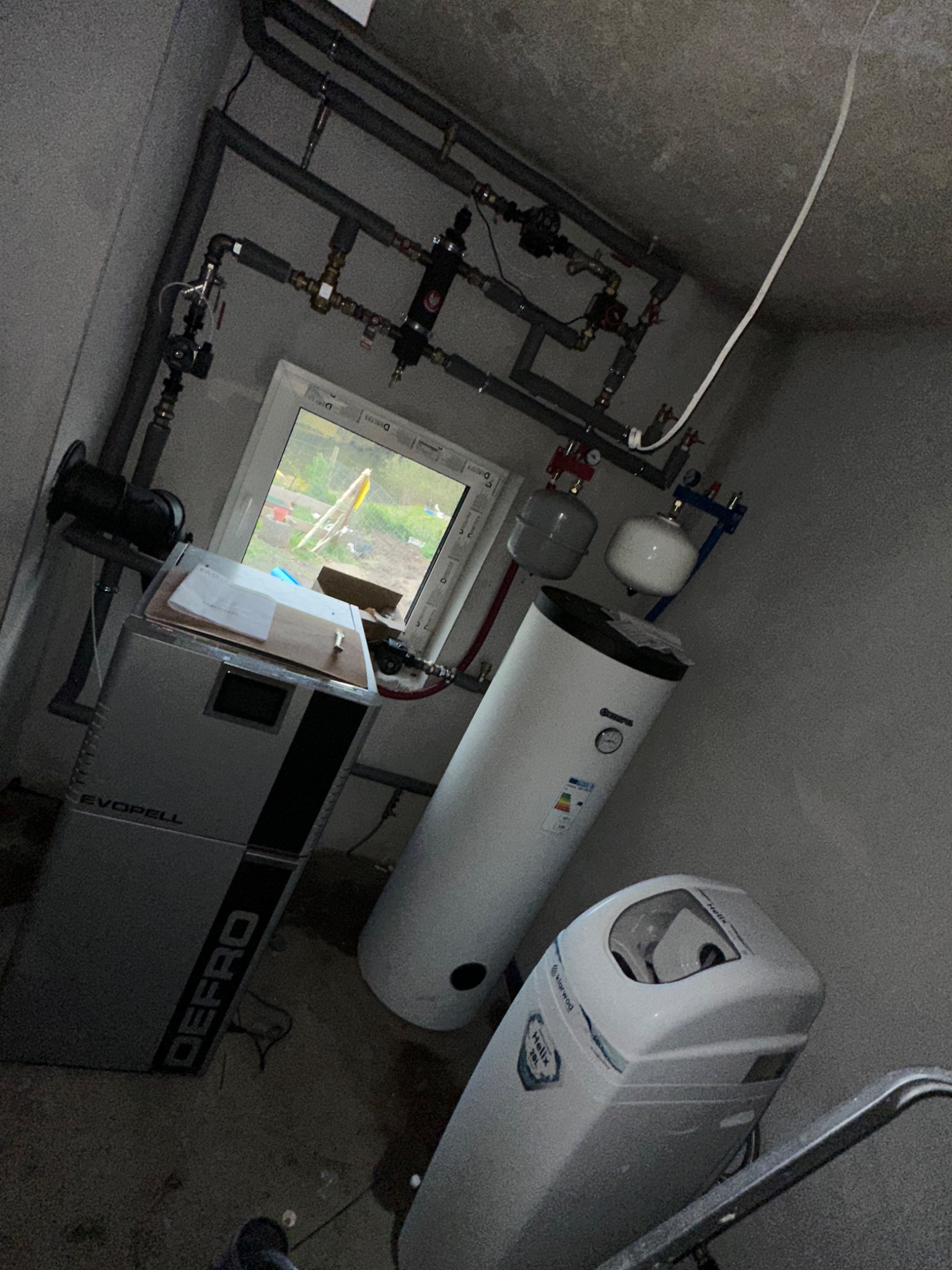 Złożona instalacja grzewcza z piecem Defro, zbiornikiem i filtrem wody w szarej kotłowni. Widoczne rury, zawory i okno z widokiem na zewnątrz.