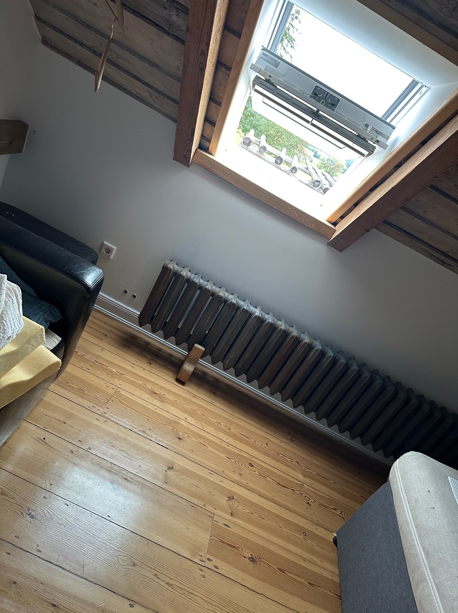 Żeliwny grzejnik żeberkowy pod oknem dachowym w pokoju na poddaszu z drewnianą podłogą i belkami stropowymi. Widoczne meble.