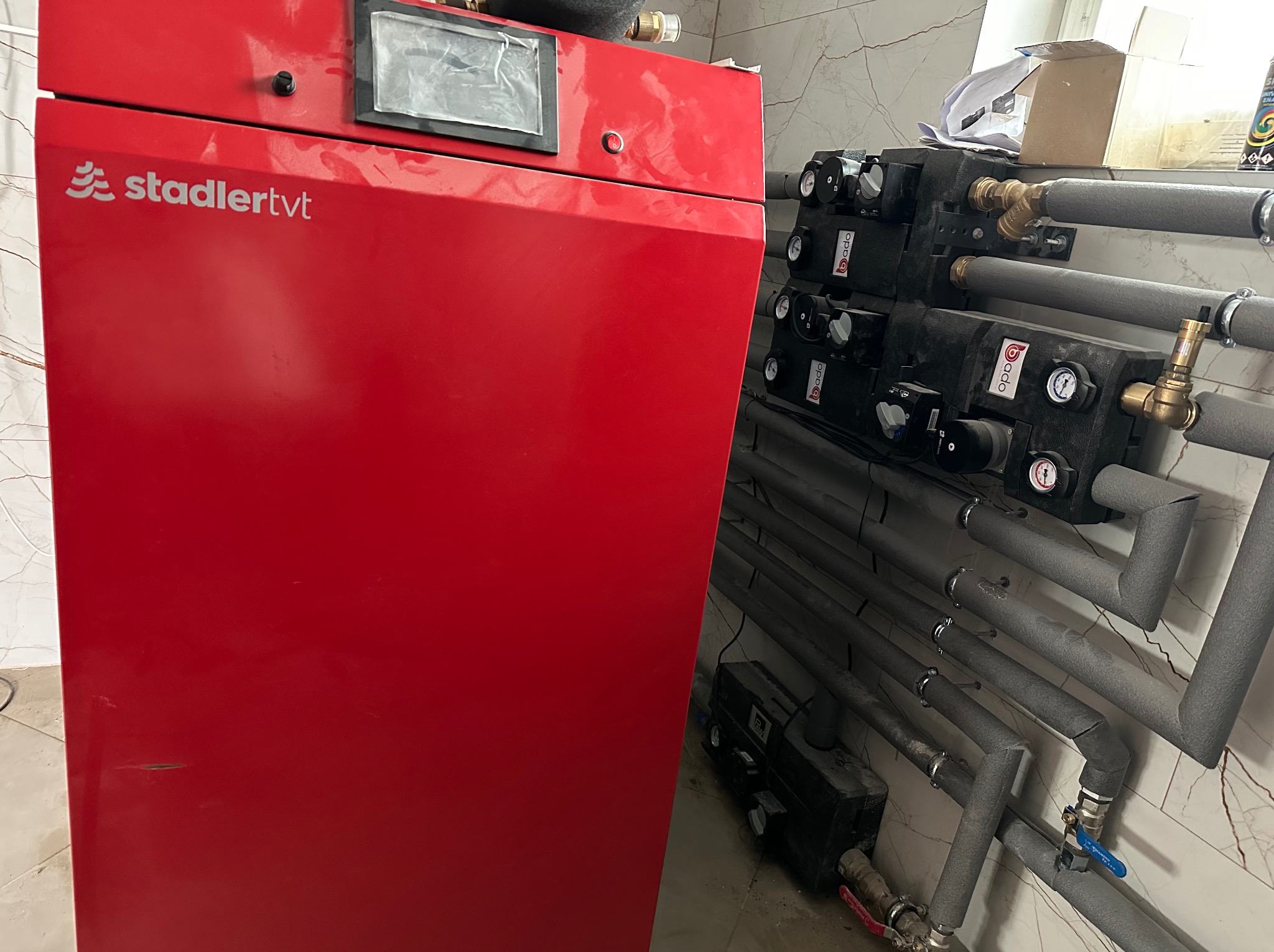 Czerwony piec Stadler TVT z czarnymi modułami i szarymi rurami w izolacji na tle marmurowej ściany. Widoczne zawory i manometry.