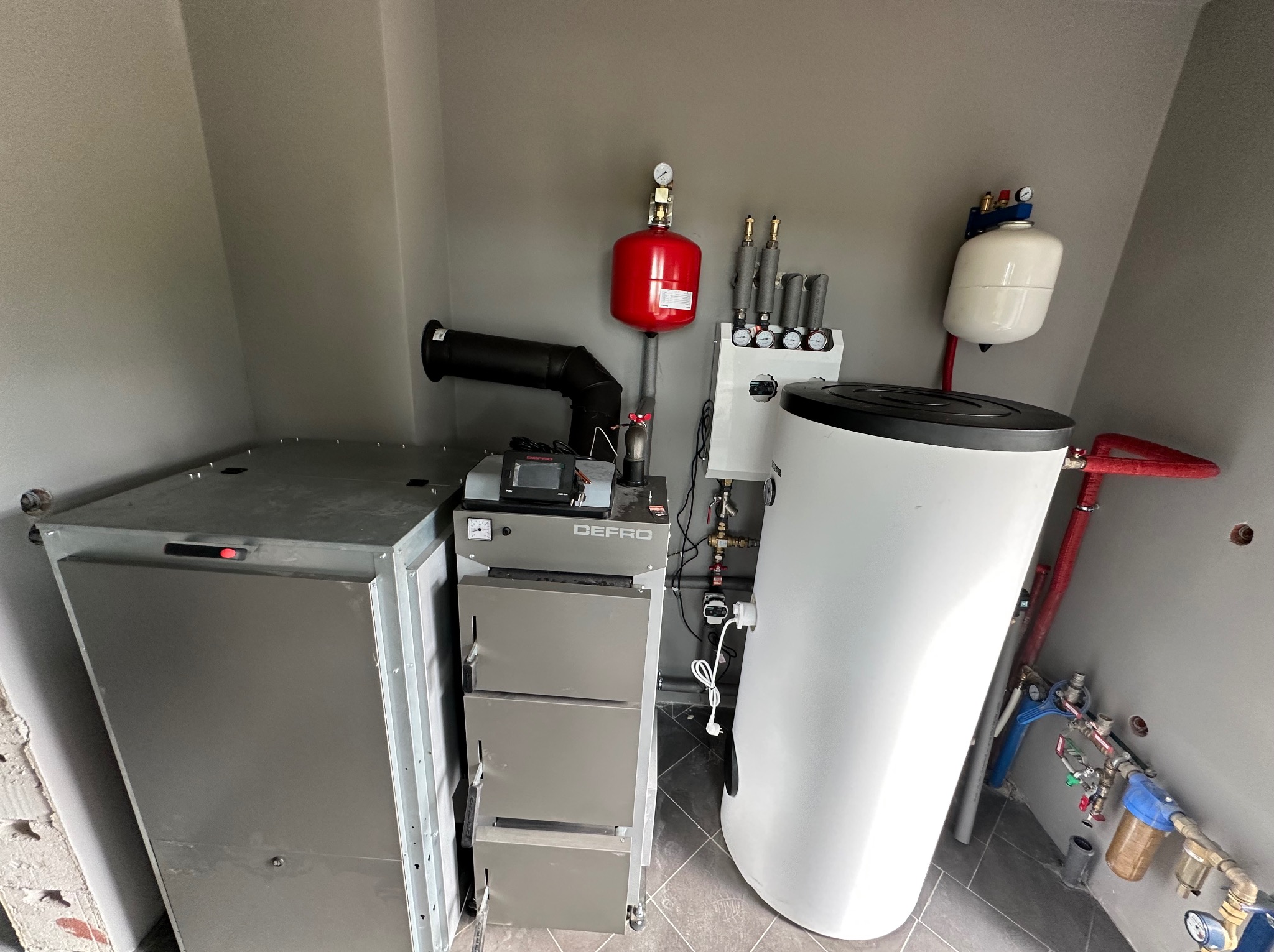 Kotłownia z piecem Defro, zbiornikiem buforowym i instalacją. Widoczne elementy hydrauliczne i grzewcze. Szare ściany i podłoga. Ujęcie z bliska.